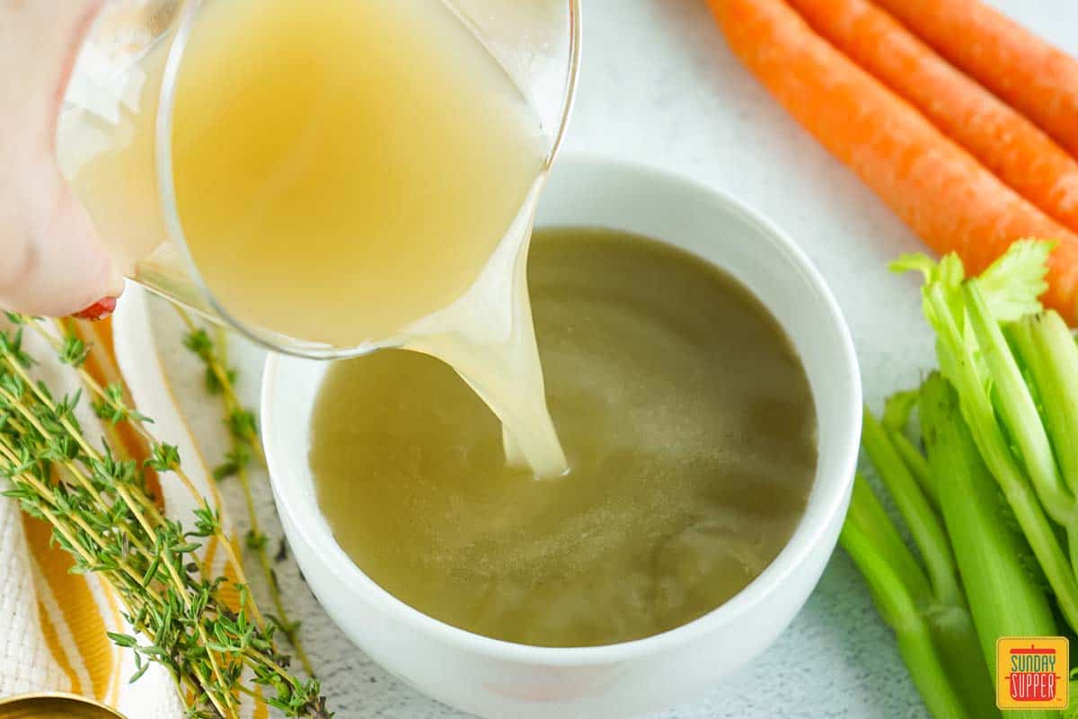 pouring turkey stock into a bowl next to celery carrots and herbs