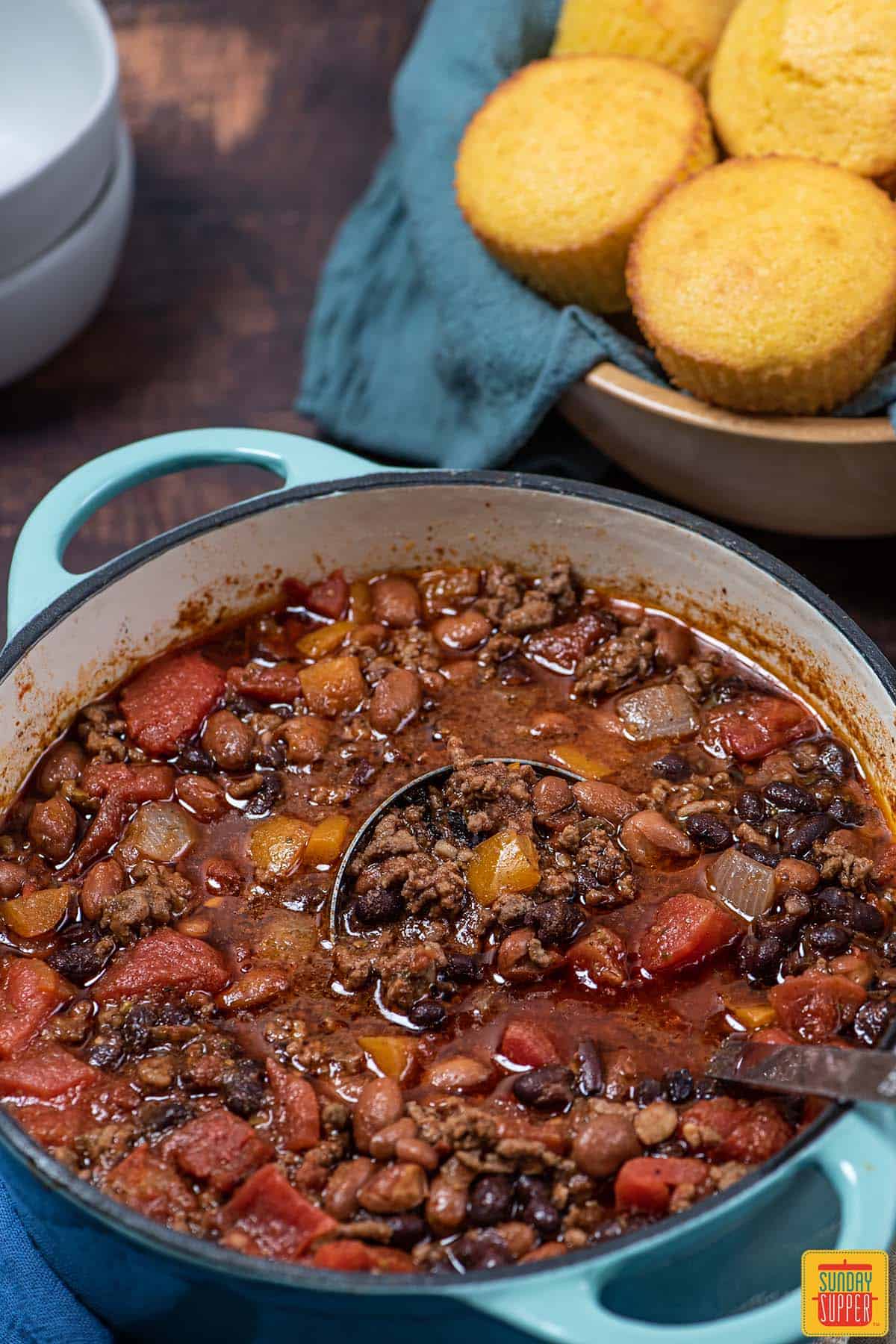 ladling beef chili in a blue pot next to cornbread