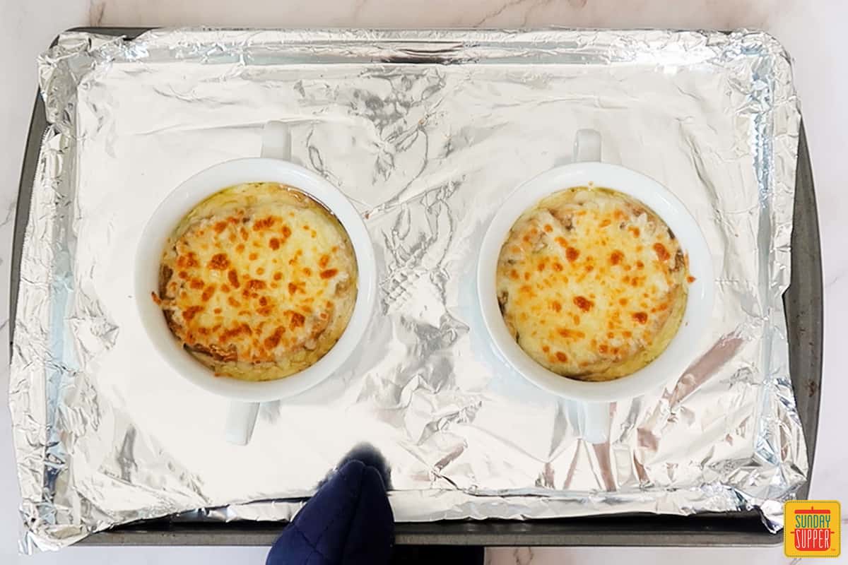 2 white bowls are French onion soup broiled on a baking sheet lined with aluminum foil