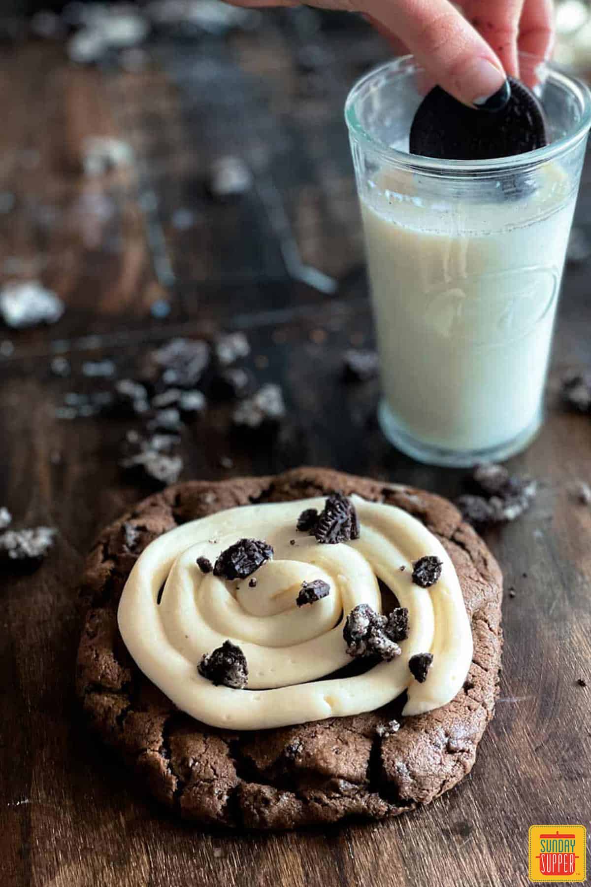 an oreo crumble cookie on a brown table with a glass of milk and an oreo being dunked into the milk