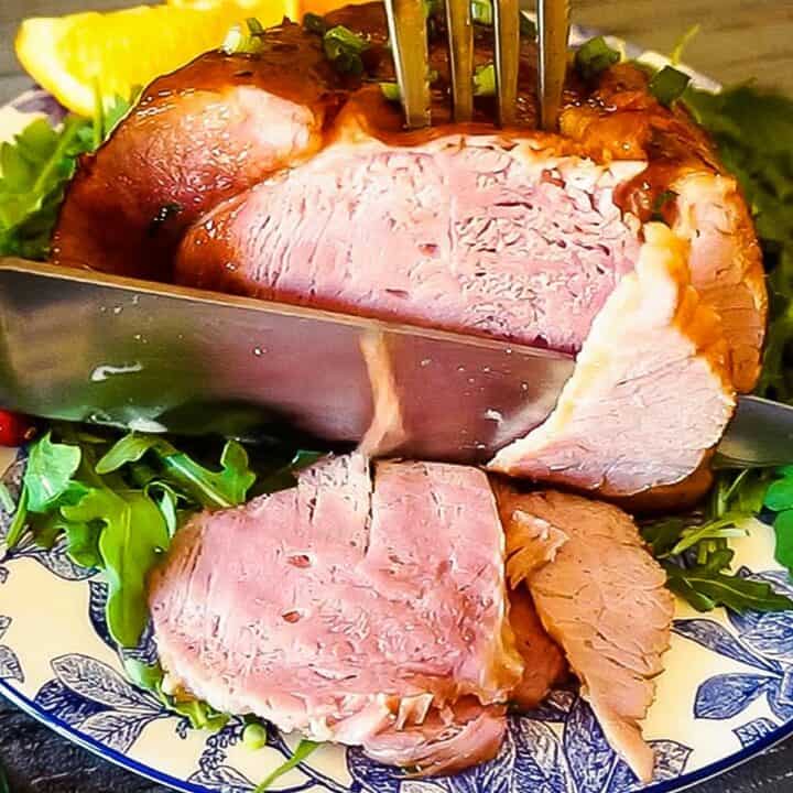 baked ham on a plate being sliced by a metal knife, with arugula and lemon