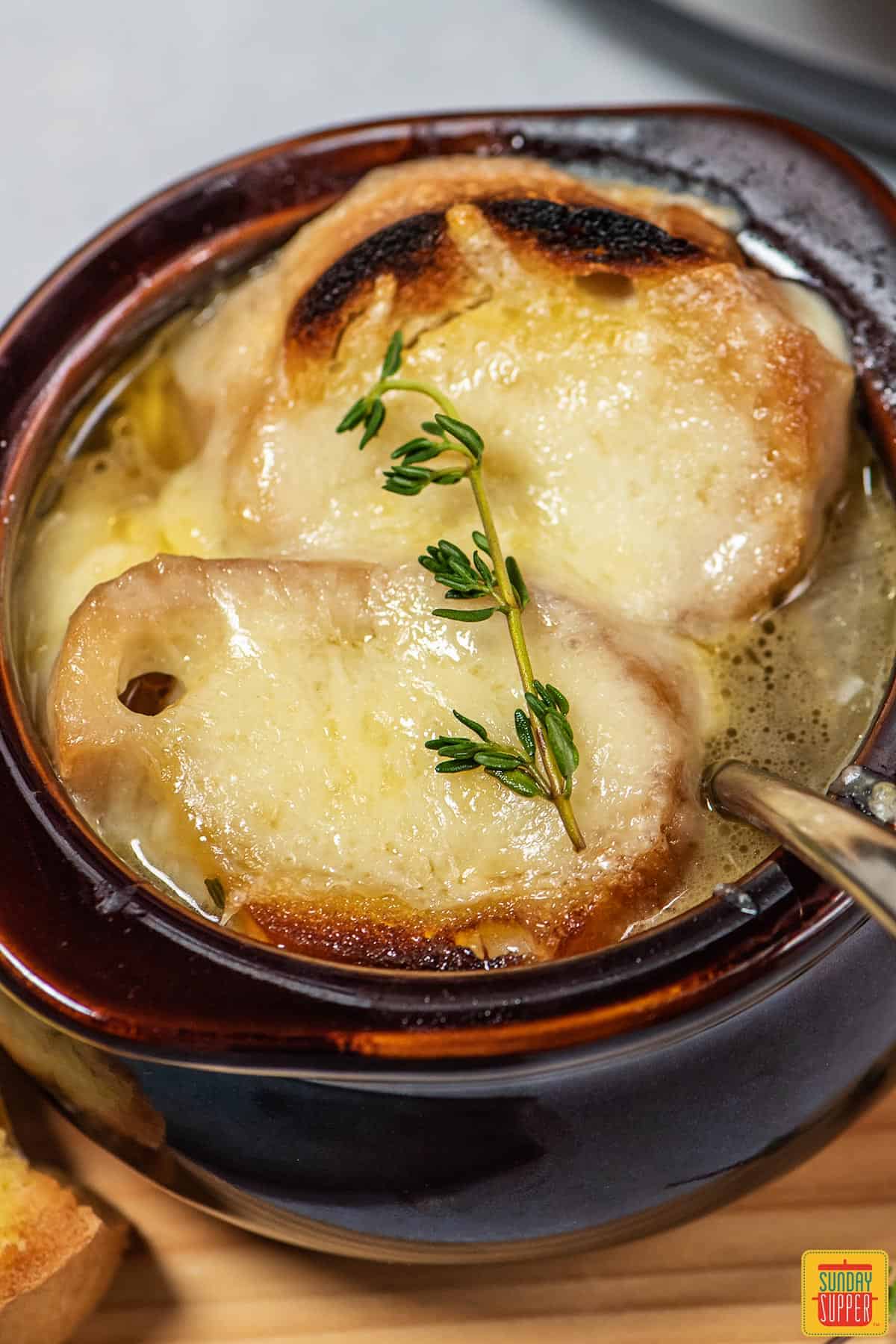 French onion soup in a brown bowl with a metal spoon inside and thyme placed on top of melty cheese
