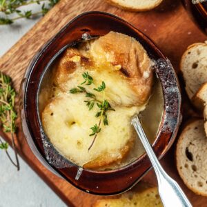 French onion soup in a brown bowl with a metal spoon inside and bread on the sides of bowl