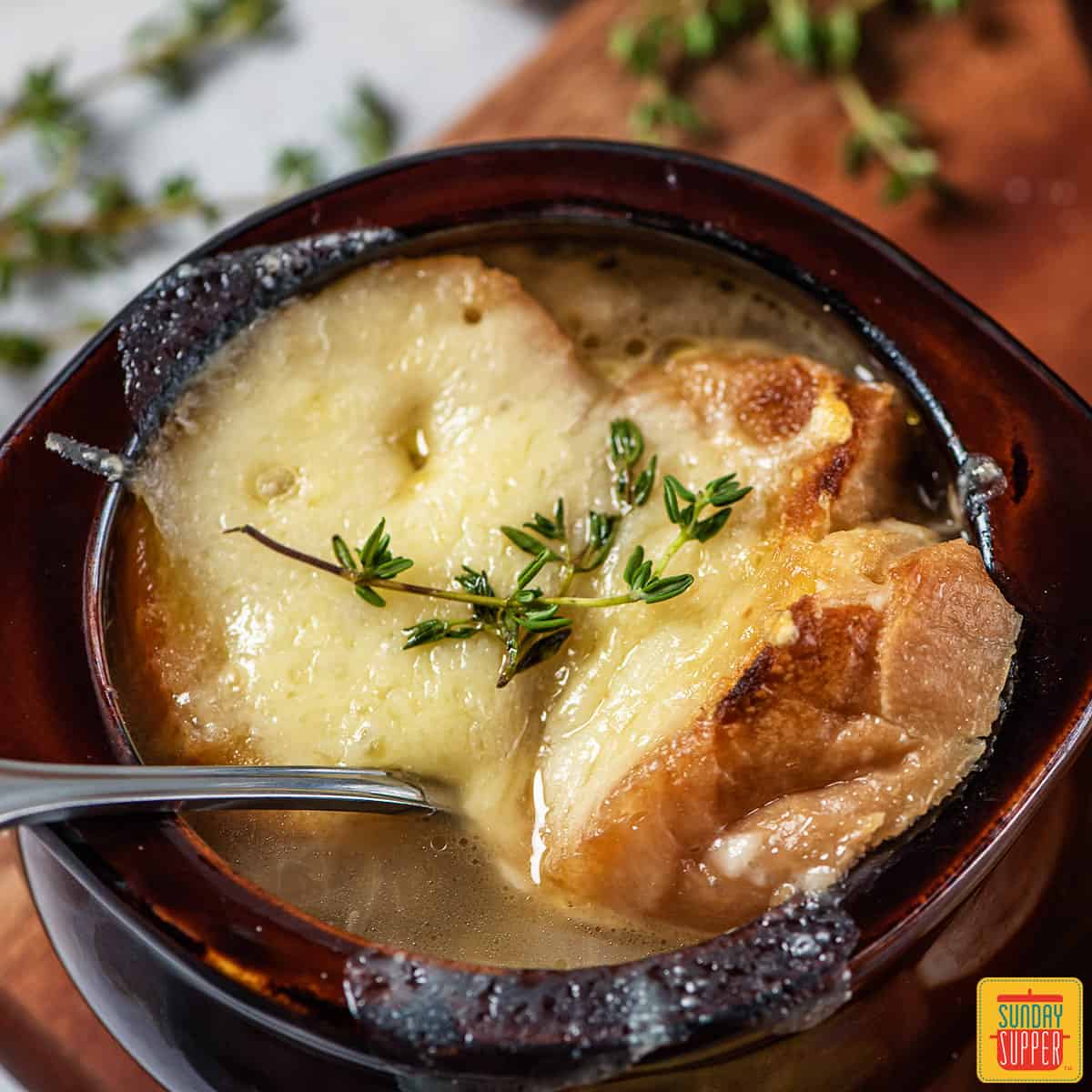 French onion soup in a brown bowl with a metal spoon inside and thyme on top