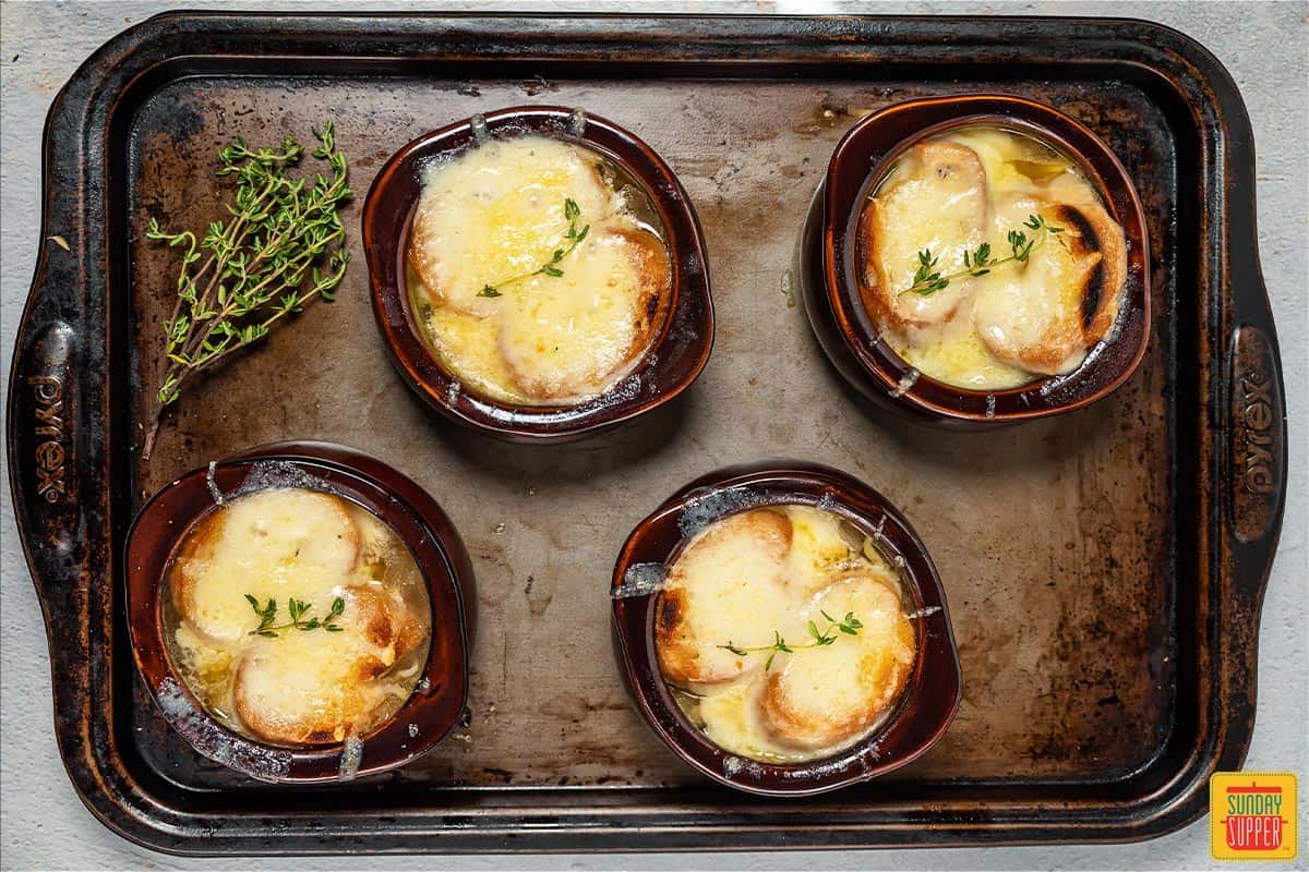 4 broiled and melty bowls of French onion soup on a baking tray