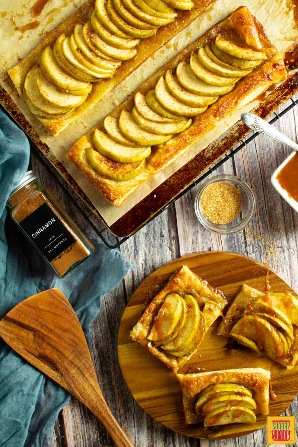 a top view of a baking sheet with two apple tarts and a cutting board with a sliced apple tart, with cinnamon, sugar and a spoon on the table