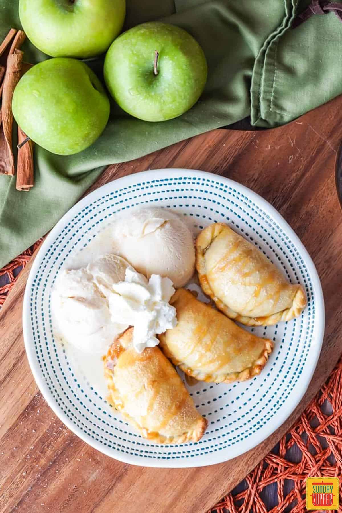 a plate of empanadas with vanilla ice cream and whipped cream on a plate next to a tea towel, green apples and cinnamon sticks
