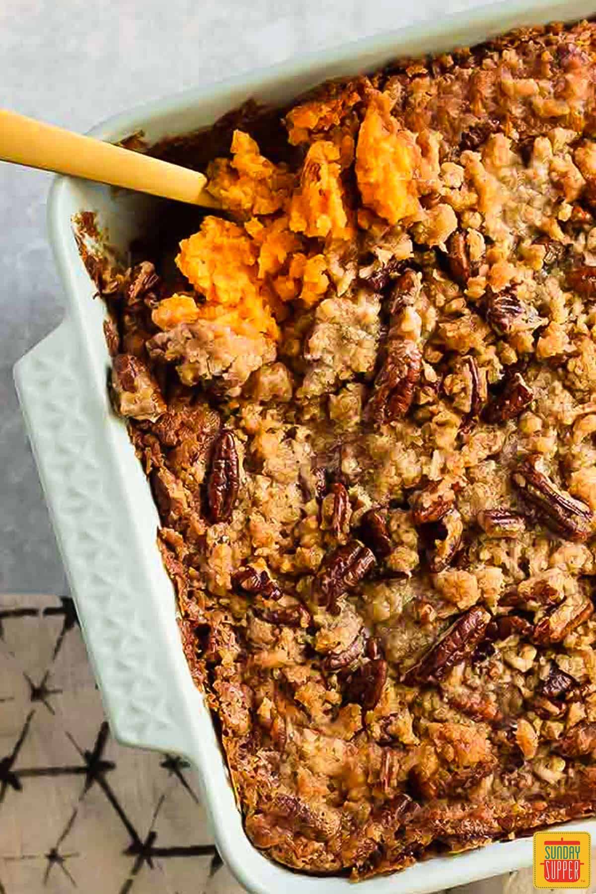 a spoon in a casserole dish of sweet potato casserole covered in pecans