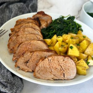 close up of sliced air fryer pork tenderloin on a white plate with roasted potatoes and sautéed spinach