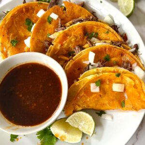 quesabirria tacos on a white plate with a white dish of birria sauce