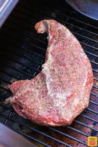 a raw tri tip steak covered in seasonings on the bars of a smoker