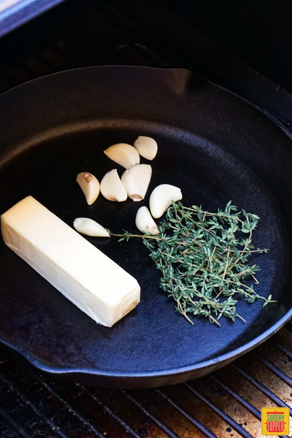 a cast iron pan in a smoker with a stick of butter, thyme sprigs, and garlic cloves