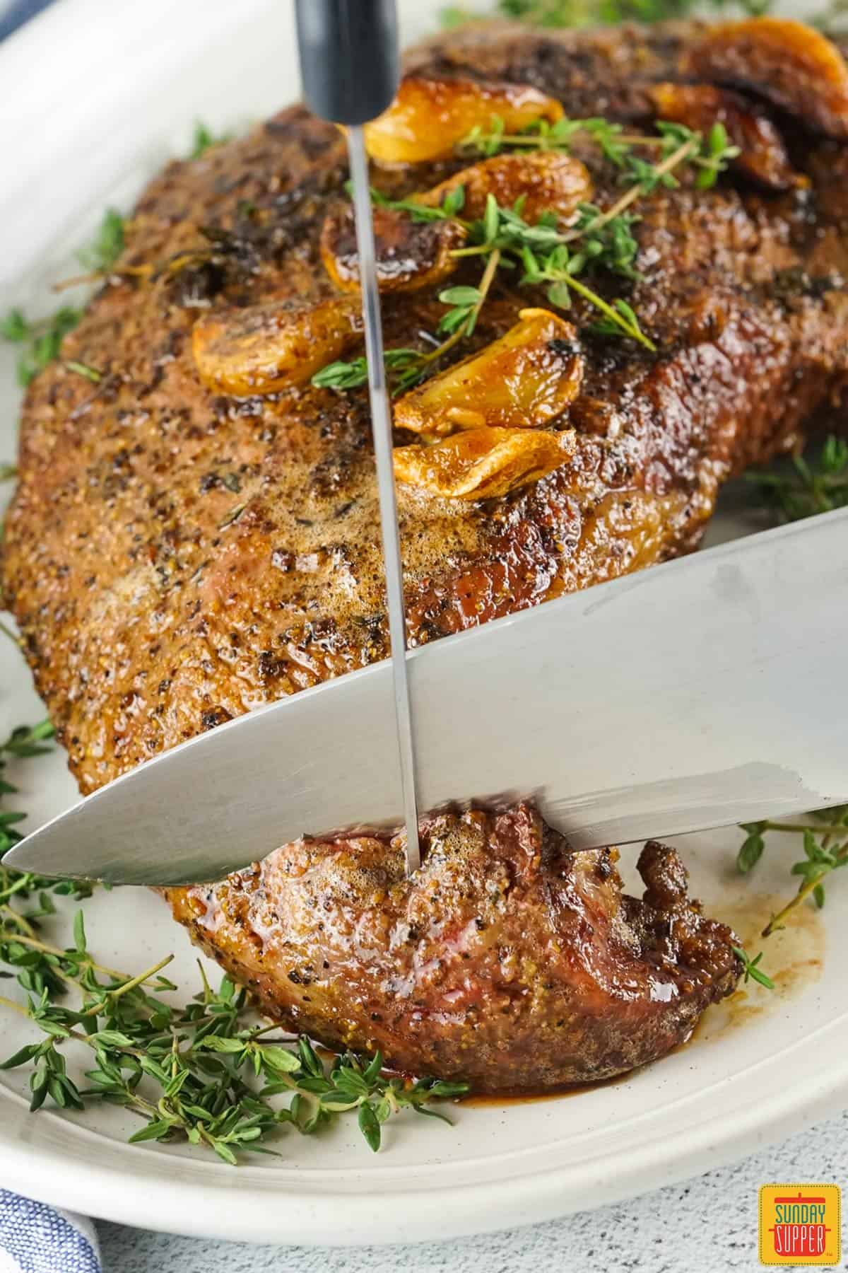 a smoked tri tip on a plate with thyme and garlic being sliced by a knife