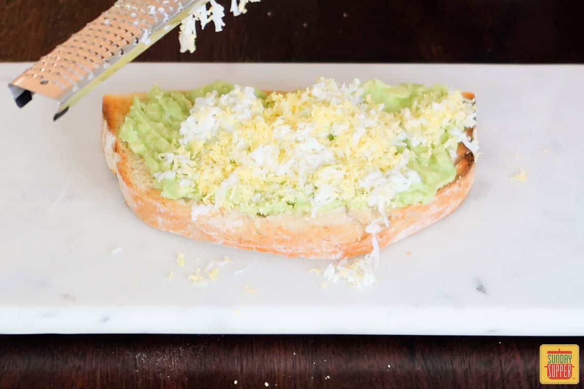 grating egg over avocado toast on a board