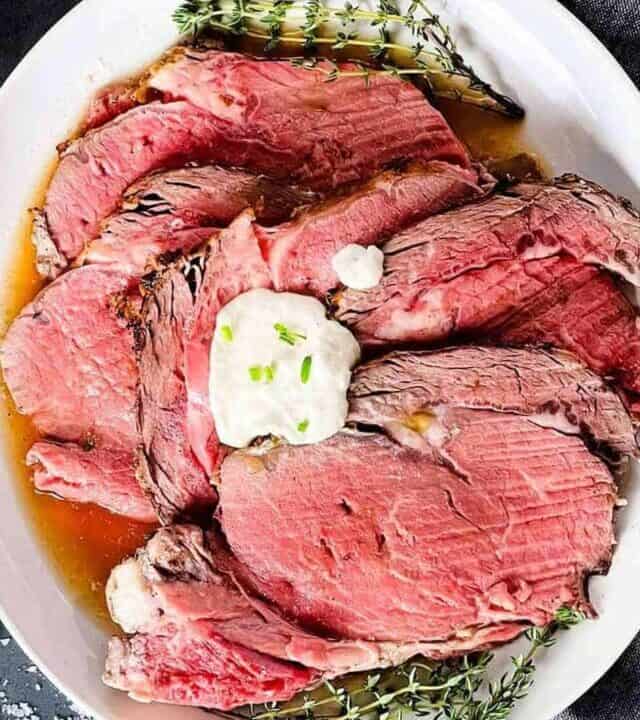 a white plate piled with sliced prime rib and au jus with a dollop of horseradish sauce