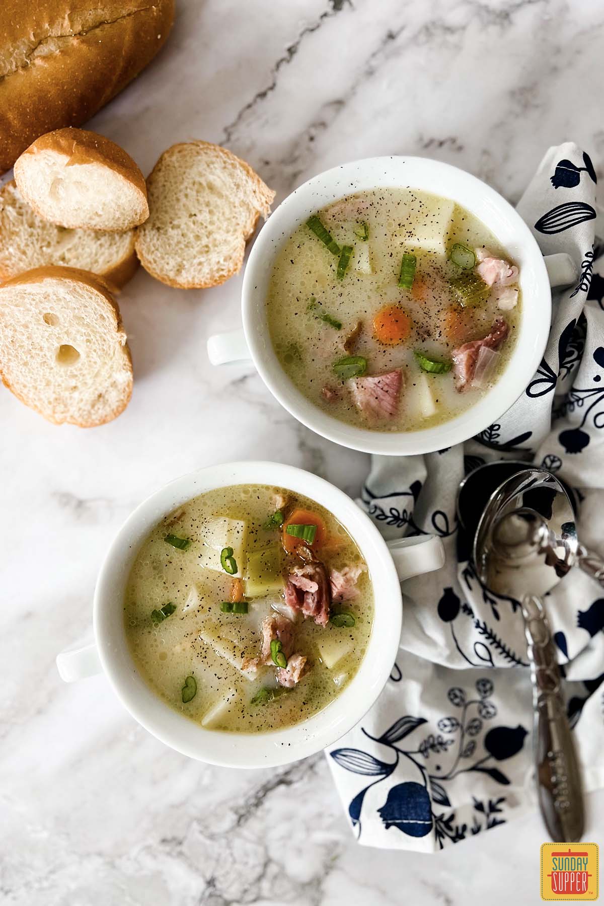 two white bowls of ham bone soup next to freshly sliced bread