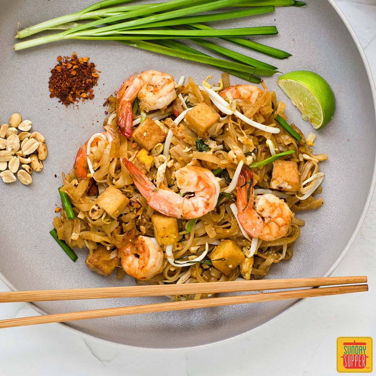 pad thai noodles on a plate with shrimp, tofu, and chopsticks to the side