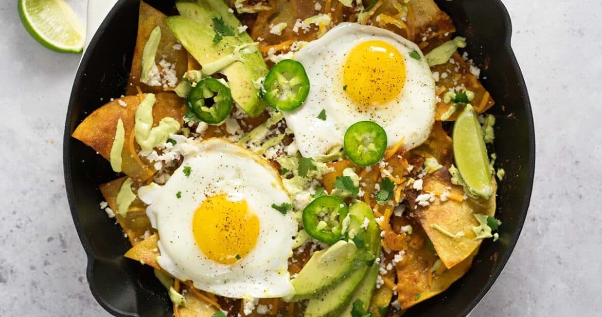 chilaquiles with fried eggs, avocado slices, jalapenos, and avocado crema in a pan