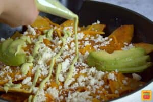adding avocado crema to chilaquiles in a pan with sliced avocados and cheese