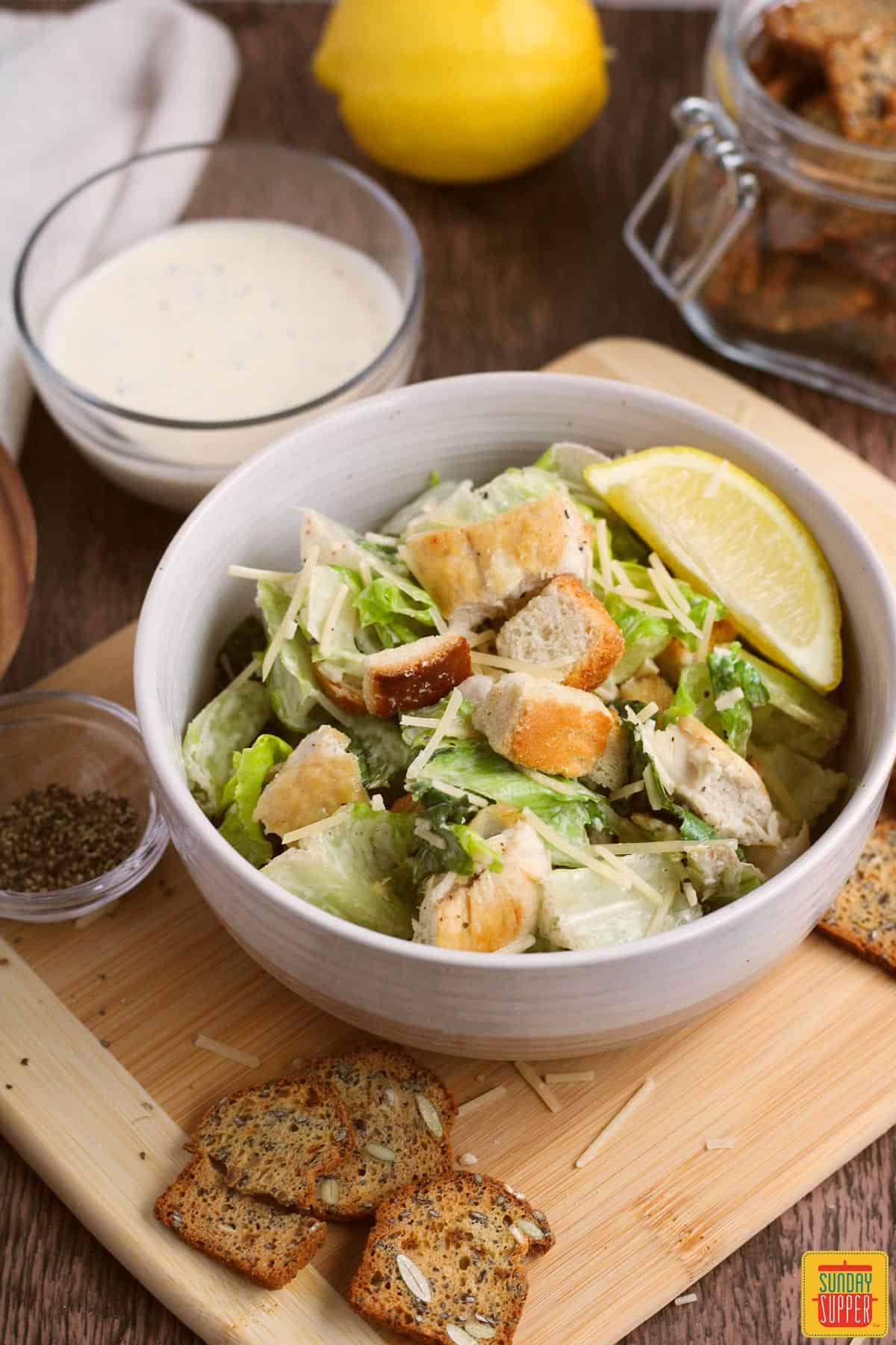 a cutting board with crouton slices, salad, pepper, dressing, and a lemon