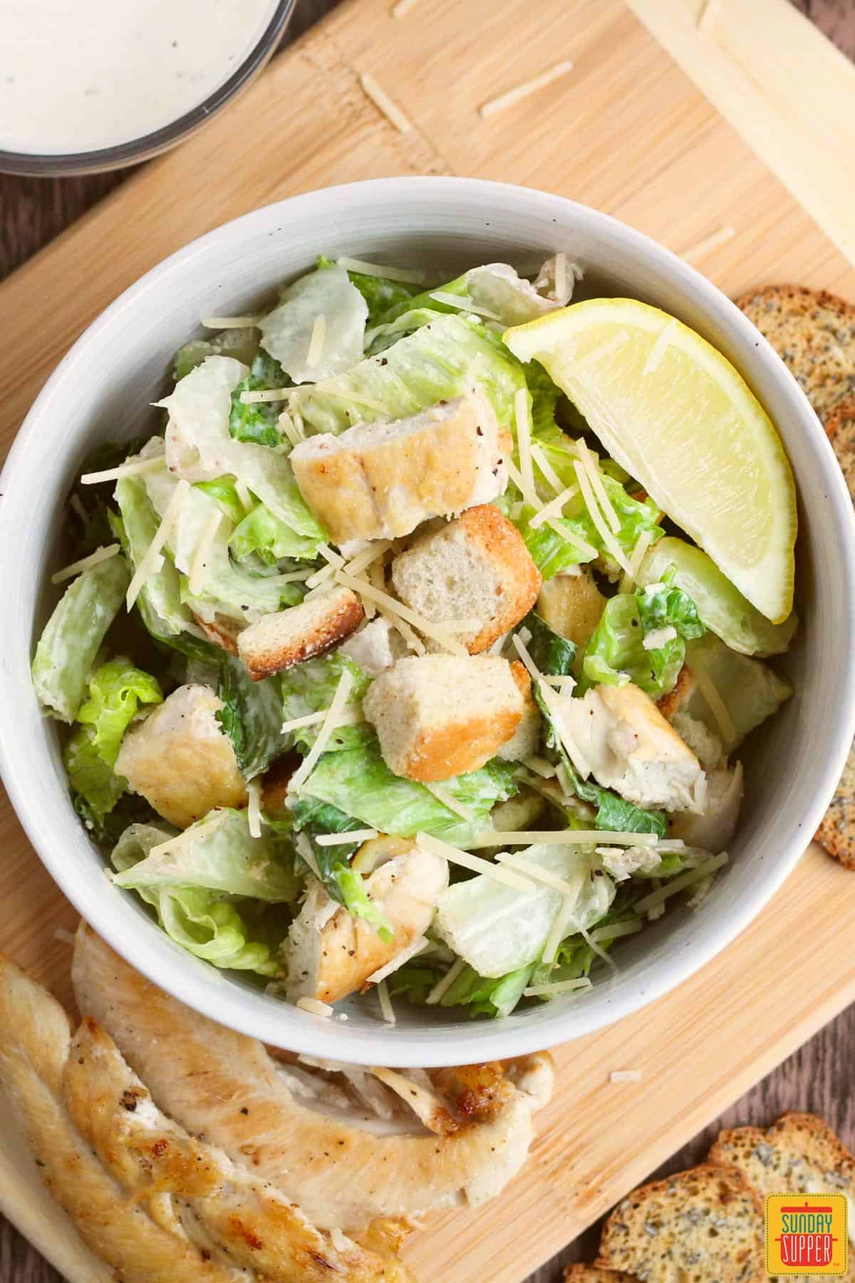 caesar salad in a bowl on a cutting board with extra grilled chicken and dressing