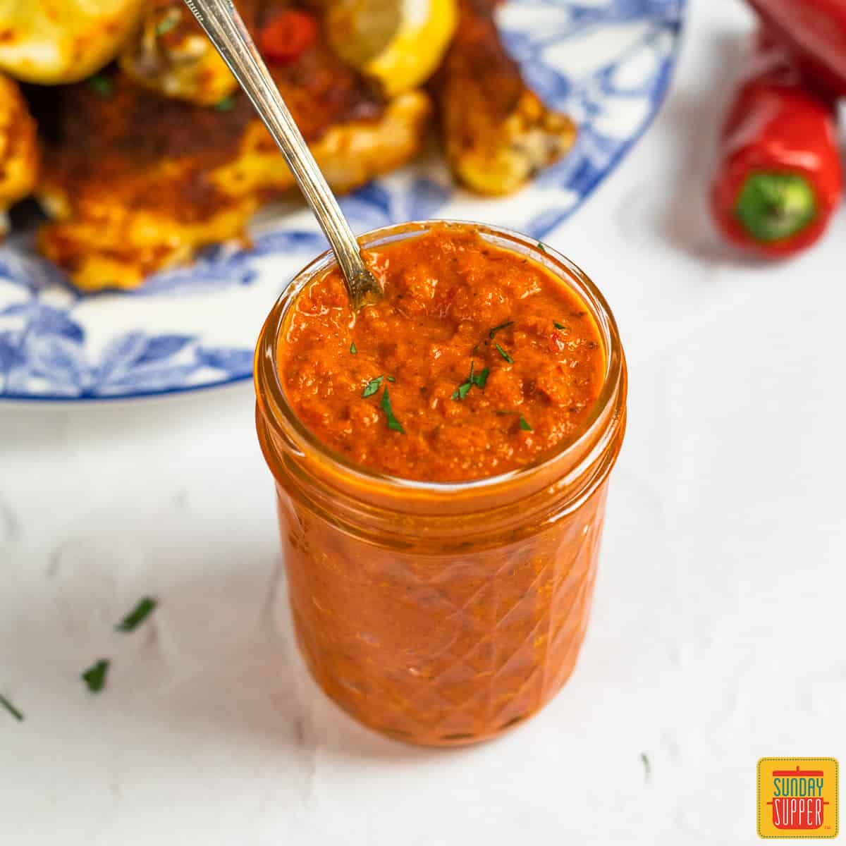 peri peri sauce in a glass jar with a spoon next to a plate of peri peri chicken