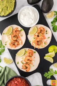 three air fryer shrimp tacos on a black platter next to limes, sour cream, avocados, salsa, and guacamole