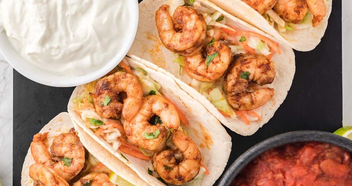 four air fryer shrimp tacos up close on a black platter next to sour cream and salsa