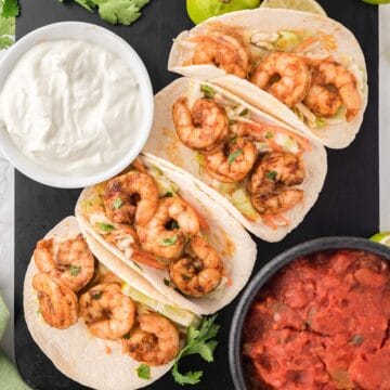 four air fryer shrimp tacos up close on a black platter next to sour cream and salsa