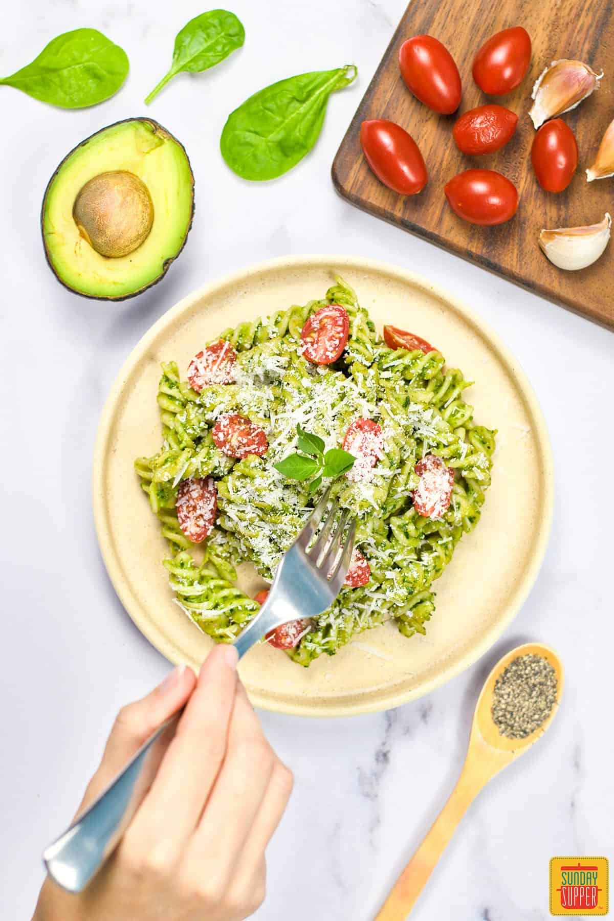 a hand taking a forkful of creamy avocado pasta off a plate with more avocado, tomatoes, pepper and garlic around