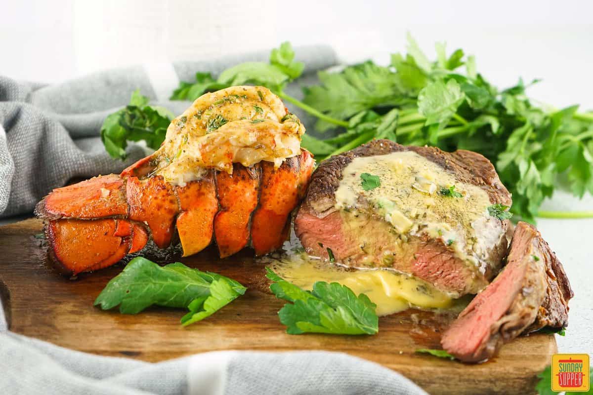 a grilled lobster tail next to grilled filet mignon on a board with herbs