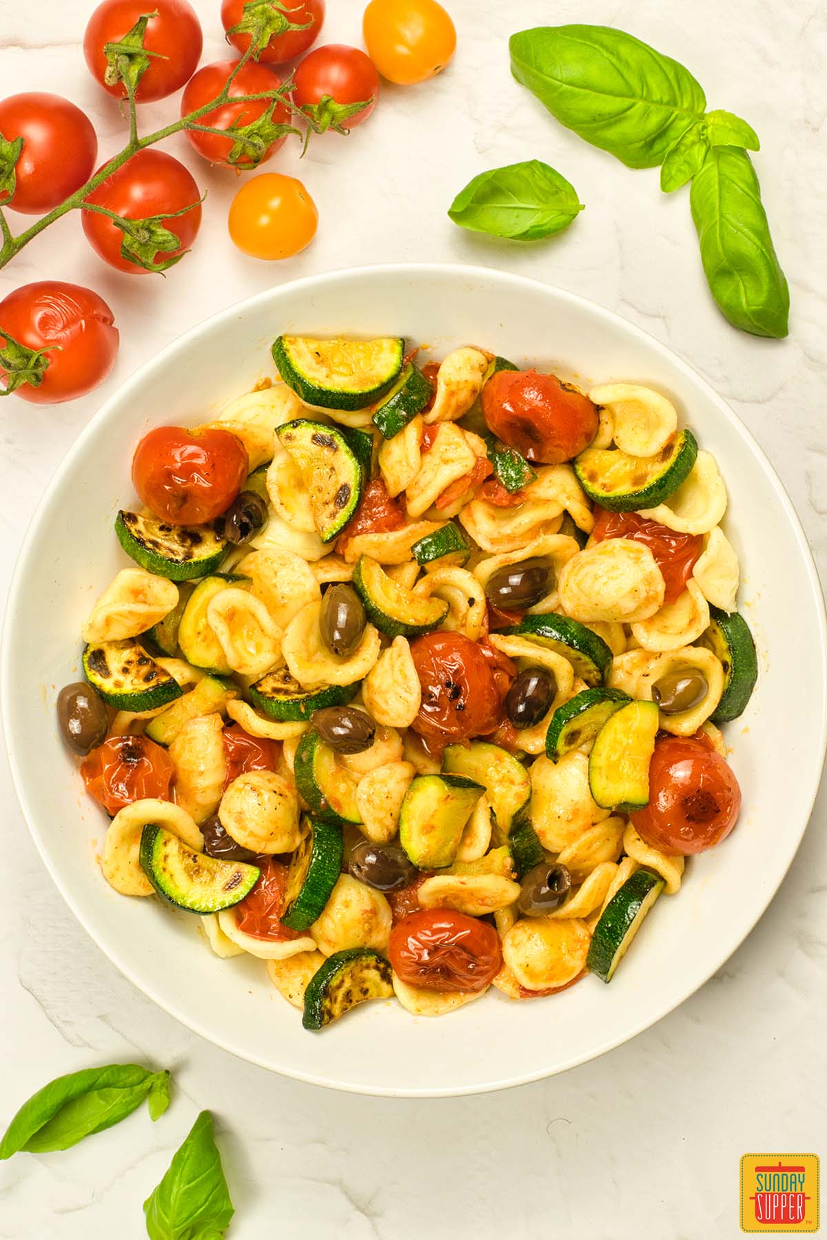 summer pasta salad in a white bowl surrounded by basil and tomatoes