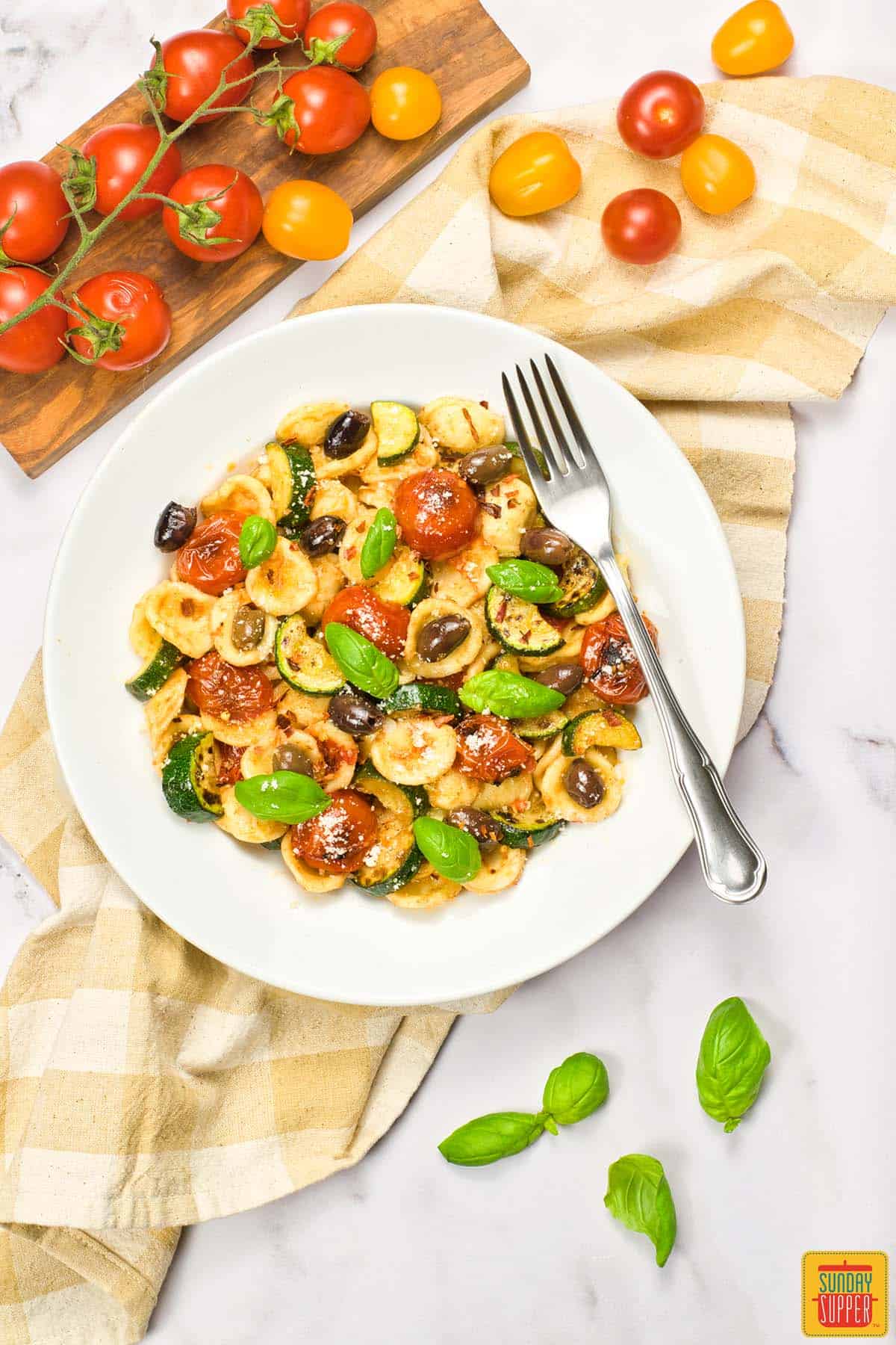 orechiette summer pasta salad on a white plate with a fork surrounded by tomatoes