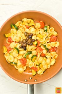 adding olives to pan with zucchini, tomatoes, and pasta
