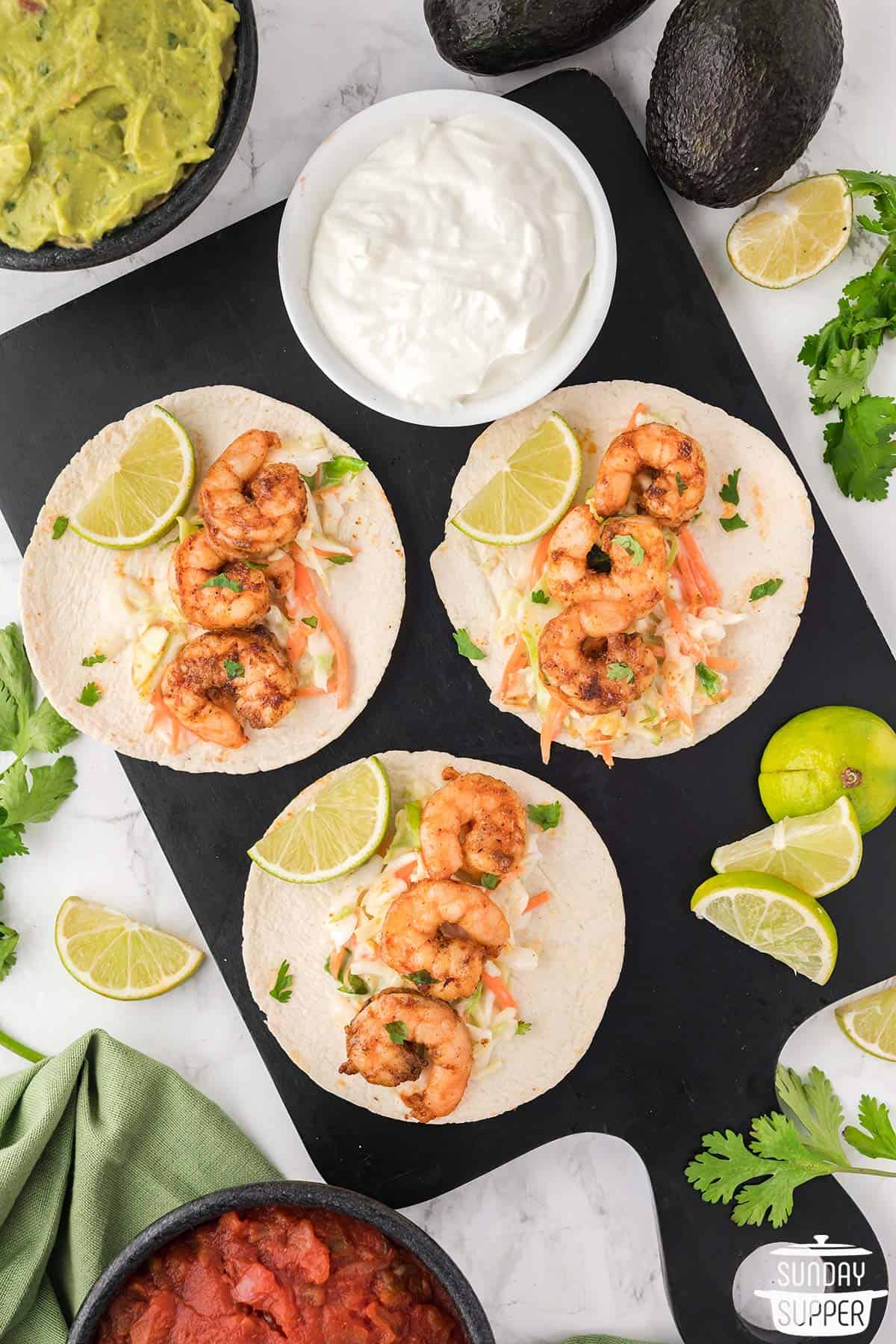 three shrimp tacos with coleslaw on a cutting board with extra toppings