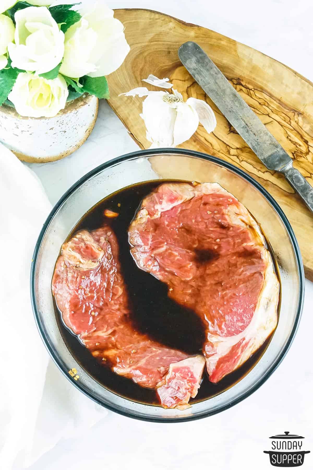 two strip steaks marinating in a bowl