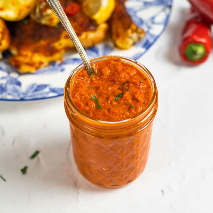 peri peri sauce in a glass jar with a spoon next to a plate of peri peri chicken