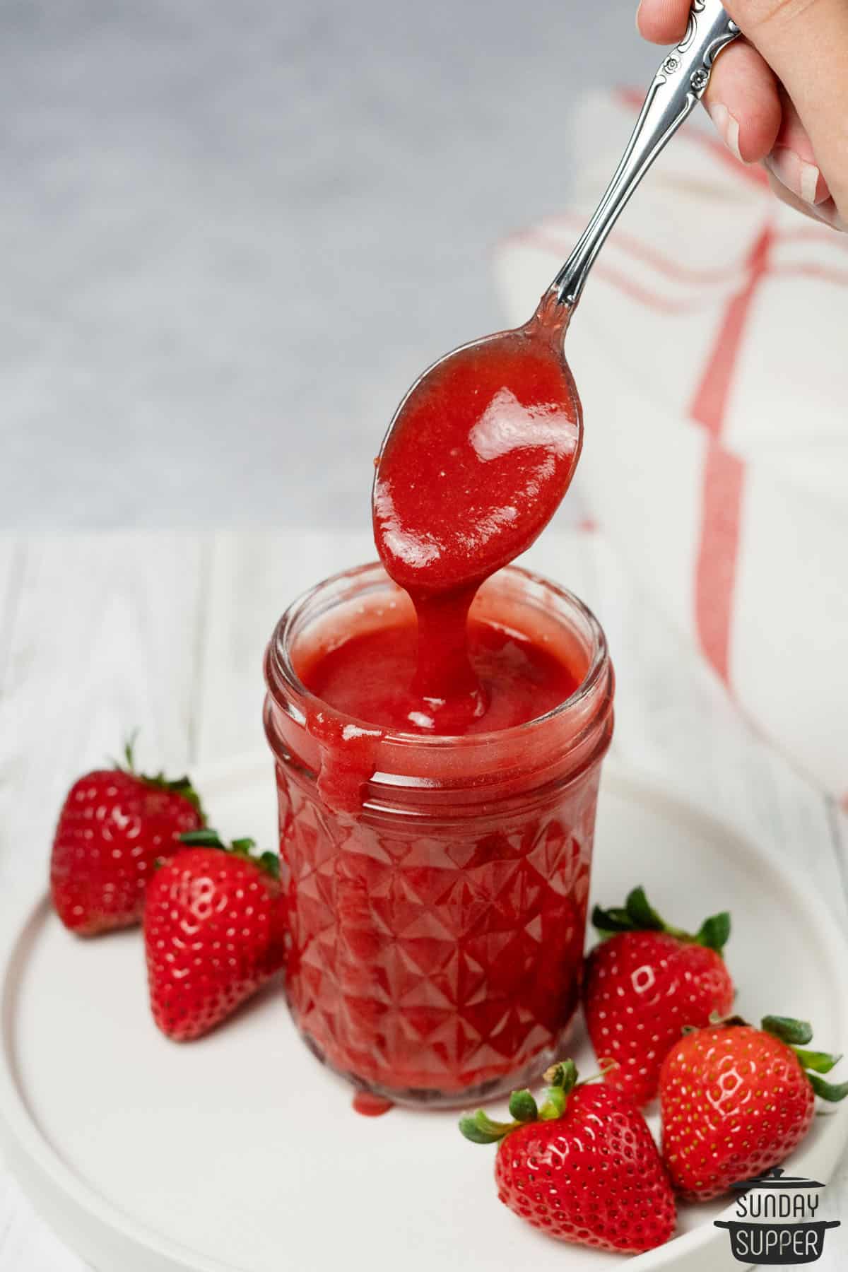 a spoon dripping strawberry sauce off in a clear mason jar with fresh strawberries on the side