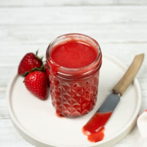 close up of strawberry sauce in a clear mason jar with fresh strawberries and a butter knife on the side