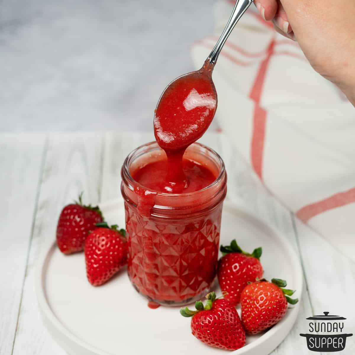 spoon inside of strawberry sauce in a clear mason jar with fresh strawberries on the side