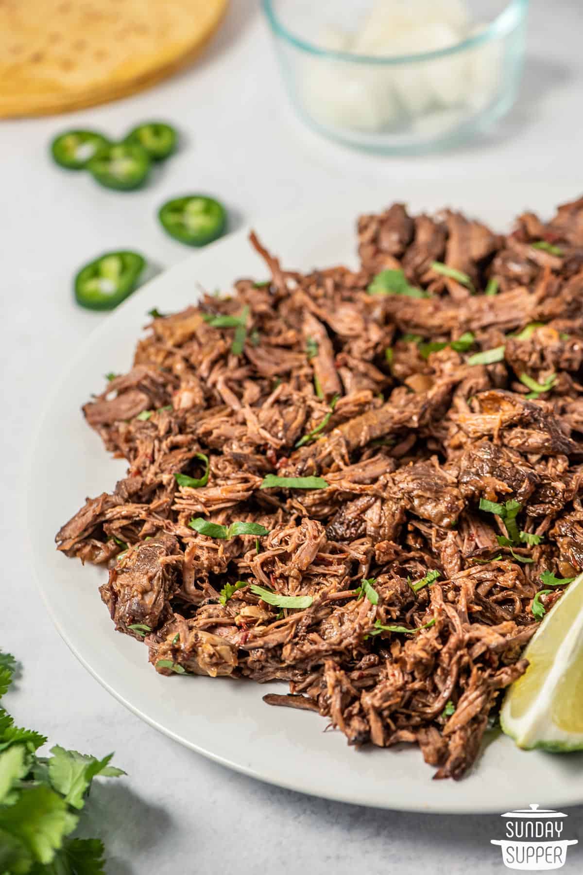 a white plate of barbacoa with jalapeno slices and lime