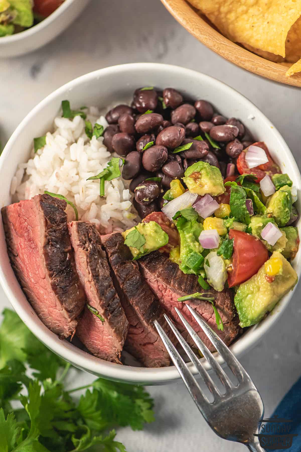 sliced flank steak in a bowl with rice, beans, and avocado salsa