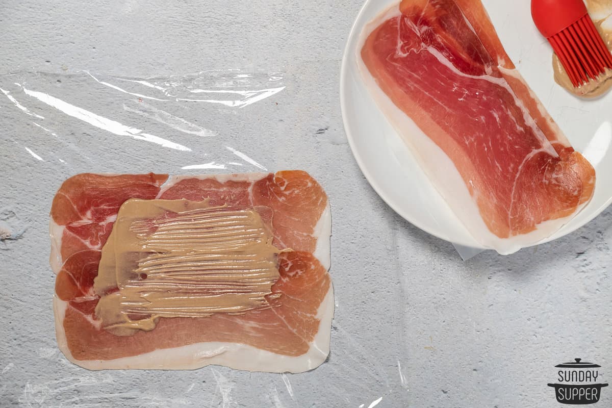 spreading dijon mustard over prosciutto on plastic wrap