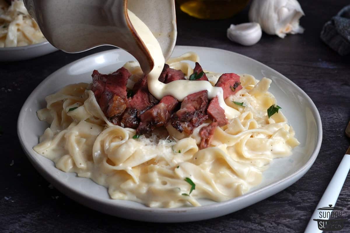 a plate of alfredo fettuccine with steak as more sauce is poured on