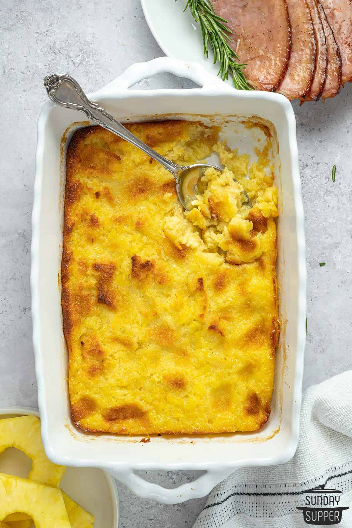 pineapple casserole in a dish with a serving spoon and slices of ham