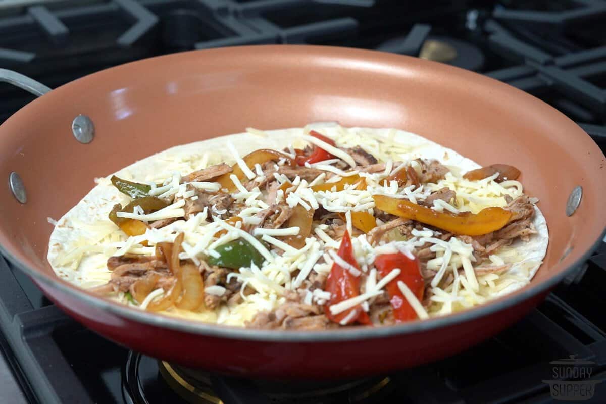 a tortilla in a pan topped with onions, peppers, pork, and cheese