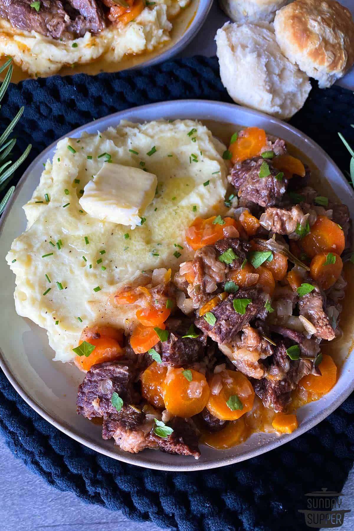 a dinner plate filled with short ribs, buttered mashed potatoes, and dinner rolls on the side