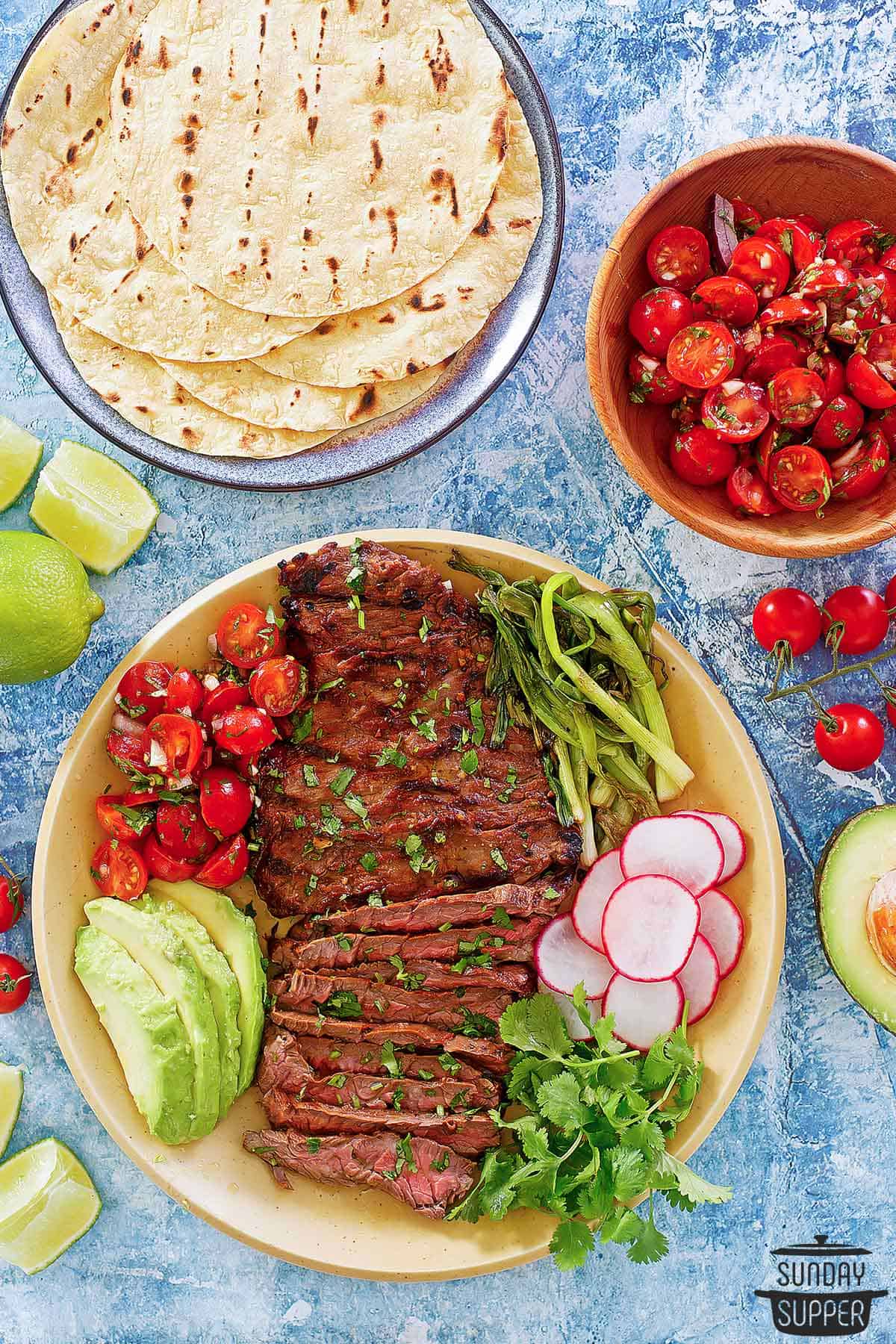 carne asada sliced on a plate with taco toppings and tortillas