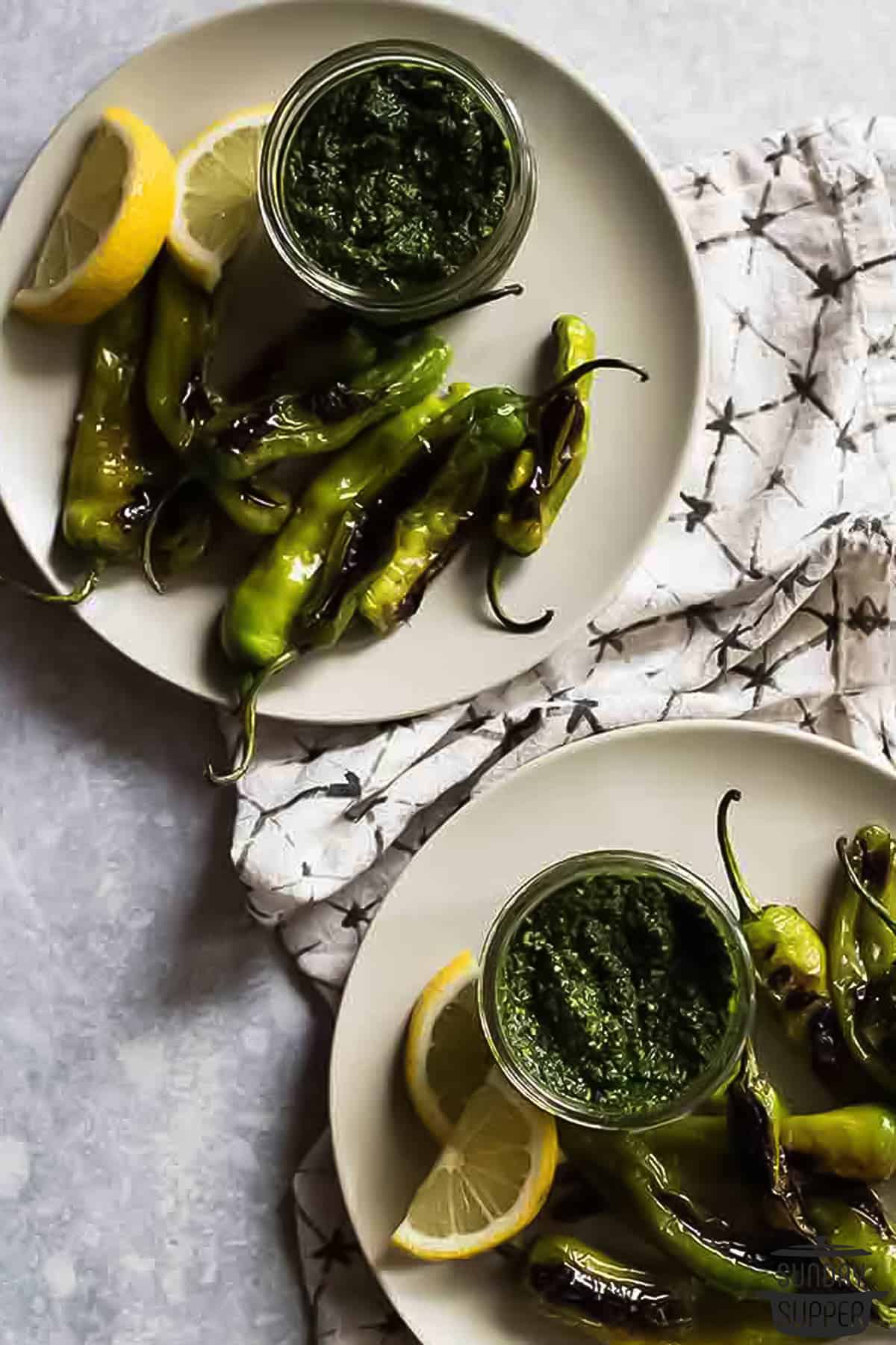 two dinner plates with blistered peppers, lemon wedges, and pesto for dipping