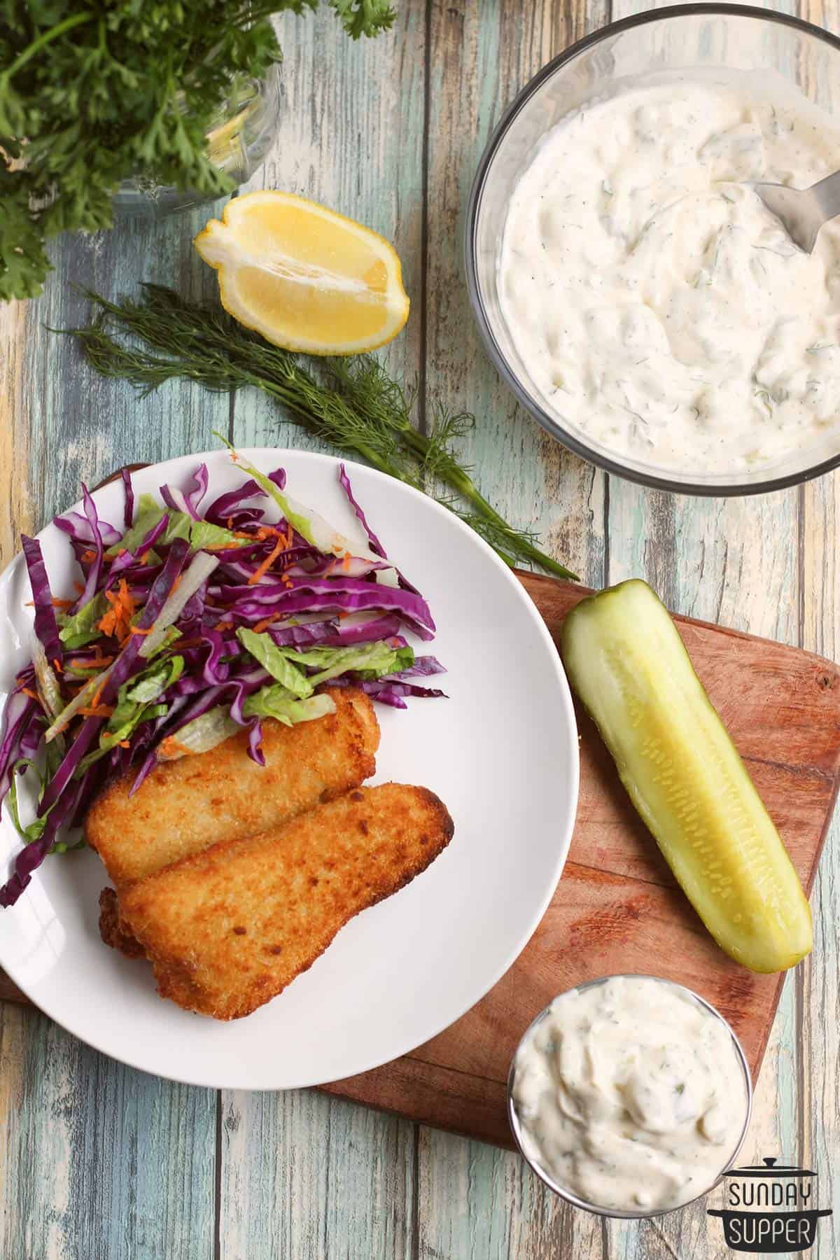 a plate of fish, coleslaw, dill pickles, and tartar sauce